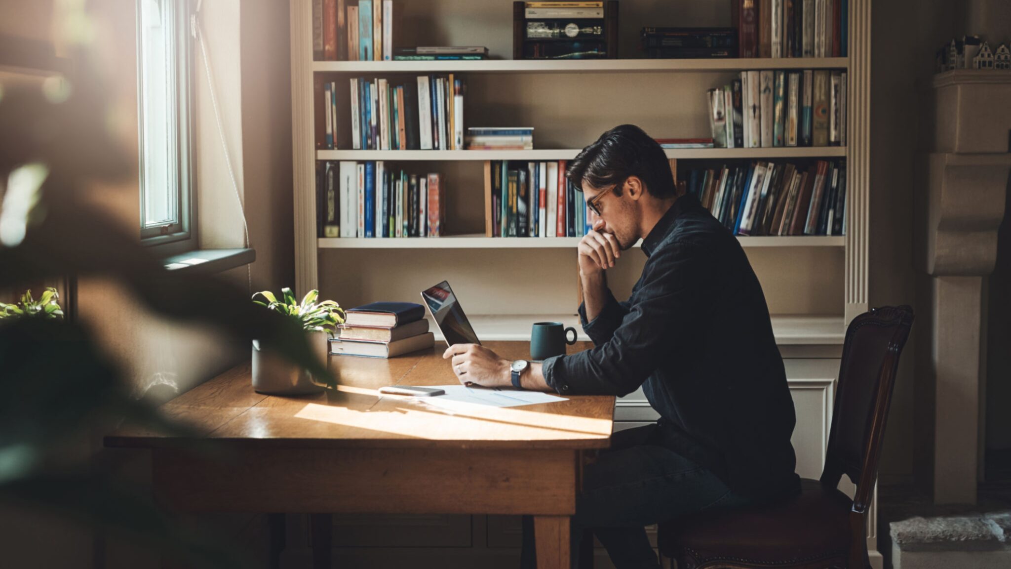 A man sits at a wooden desk in a home library, thoughtfully looking at a tablet while resting his chin on his hand.