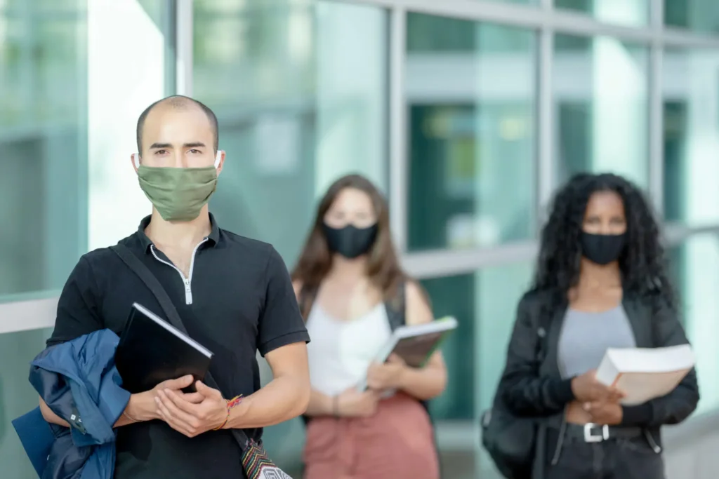 Four diverse students wearing face masks, holding books and folders.